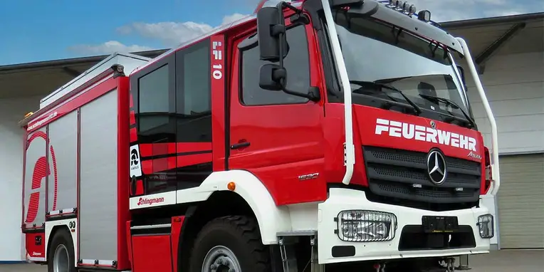 Rotes Feuerwehrfahrzeug von Mercedes-Benz mit Aufschrift „FEUERWEHR“ an der Front, steht vor einer Gebäudewand. Das Fahrzeug ist mit umfangreicher Ausstattung für den Einsatz versehen.
