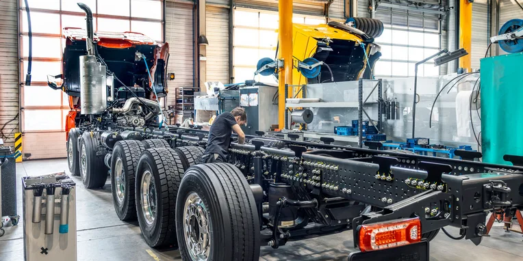 Meccanico che lavora su un telaio di camion Mercedes-Benz in un'officina, con due cabine di camion visibili sullo sfondo.