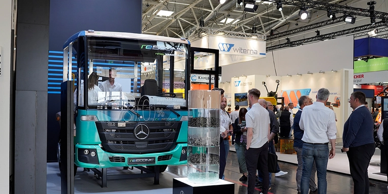 Ein Mercedes-Benz Bus steht an einem Messestand, während Besucher den Bus betrachten und sich unterhalten. Im Hintergrund sind verschiedene Stände und Aussteller auf der Messe zu sehen.