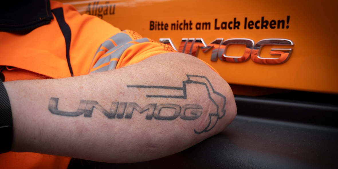 Tattoo eines UNIMOG-Logos auf dem Unterarm einer Person, neben einem orangefarbenen UNIMOG-Fahrzeug mit einem humorvollen Hinweis, nicht am Lack zu lecken.