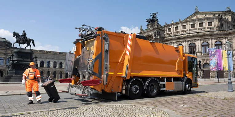 Ein orangefarbener Mercedes-Benz Econic Müllwagen steht auf einem Platz, während ein Arbeiter in orangefarbener Uniform eine Mülltonne leert.