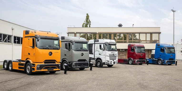 Cinquanta camion Mercedes-Benz in fila, assortiti nei colori arancione, grigio, bianco, rosso e blu, esposti in un'area di parcheggio.