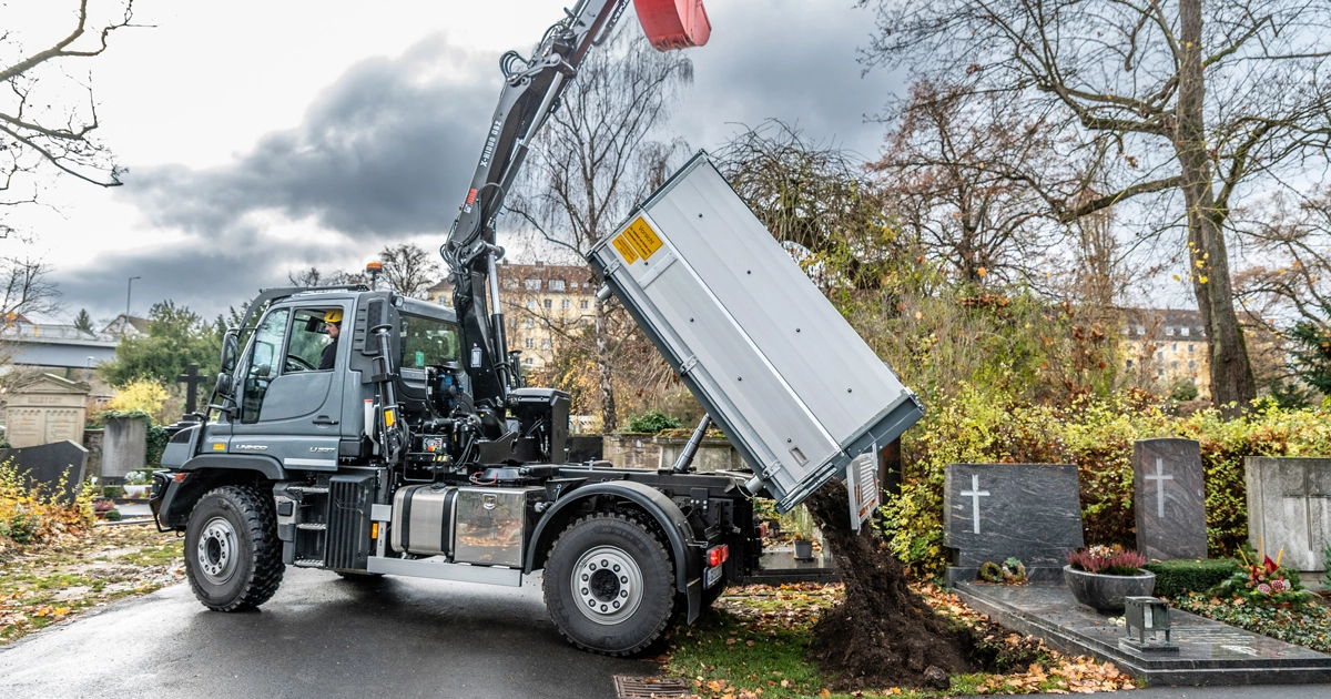 Ein Mercedes-Benz-Lkw hebt eine Ladeschaufel mit Erde an, während er auf einem Friedhof steht. Im Hintergrund sind Grabsteine und Herbstlaub sichtbar.