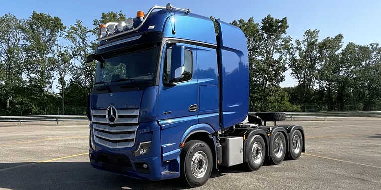 Camion Mercedes-Benz di colore blu, con cabina alta e quattro assi, parcheggiato su un’area asfaltata, circondato da alberi sullo sfondo.