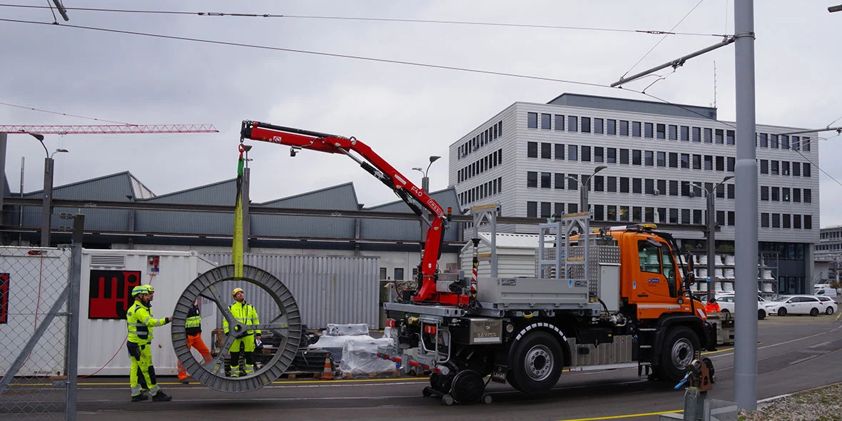 Ein Mercedes-Benz Unimog hebt mit einem Kran eine große Kabelrolle an. Zwei Arbeiter in Sicherheitskleidung stehen in der Nähe und überwachen den Vorgang auf einer Baustelle.