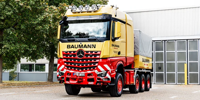 Camion Mercedes-Benz con livrea gialla e rossa, marchiato Baumann, parcheggiato davanti a un edificio. Presenta fari sopra cabina e tre assi posteriori.