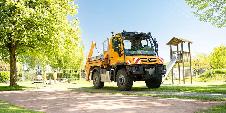 Ein gelber Mercedes-Benz Lkw steht auf einem gepflasterten Weg in einem Park, umgeben von Bäumen und einer Spielplatzstruktur im Hintergrund.