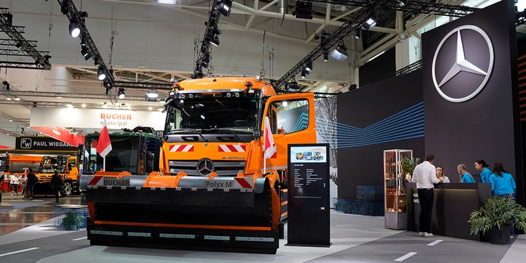 Ein orangefarbener Mercedes-Benz Lkw mit einem Schneepflug steht in einem Ausstellungsgelände. Im Hintergrund sind Menschen mit Informationsständen und Banner zu sehen.