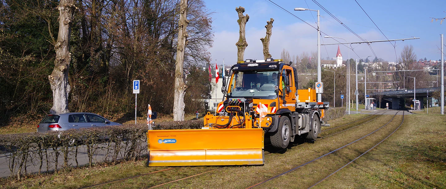 Ein orangefarbener Schneepflug von Mercedes-Benz steht auf der Gleisbettfläche neben einer Strasse, bereit zum Einsatz. Im Hintergrund sind einige Bäume und eine Haltestelle sichtbar.