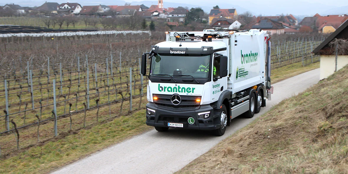 Ein Müllfahrzeug von Mercedes-Benz fährt auf einem Feldweg zwischen Weinreben. Im Hintergrund sind moderne Gebäude und eine eher ländliche Landschaft zu sehen.