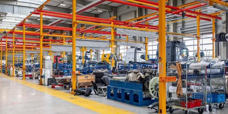 Linee di assemblaggio in una fabbrica Mercedes-Benz, con veicoli commerciali e motori visibili. I colori vivaci delle strutture portanti evidenziano l'ambiente industriale.