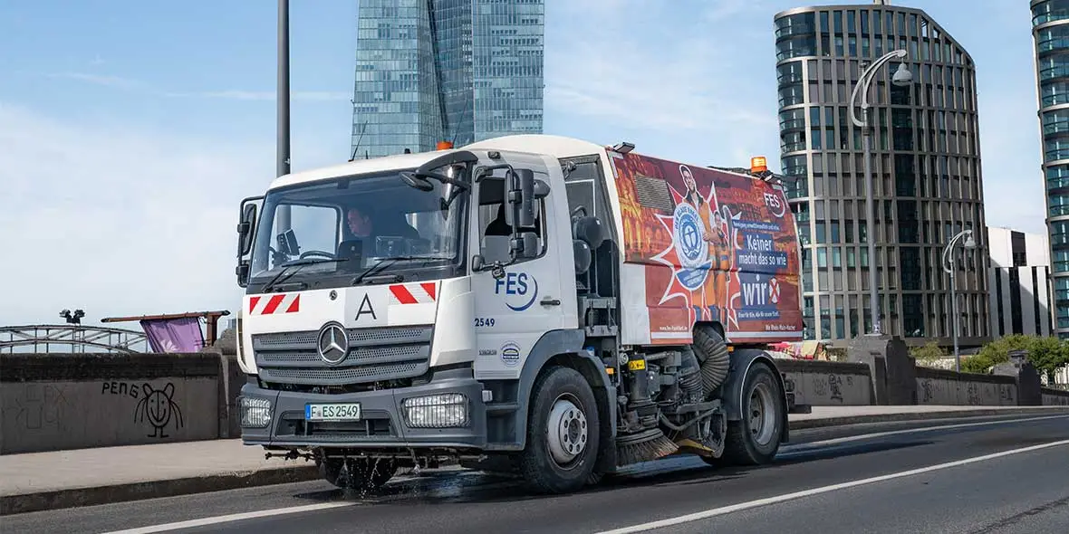 Fahrzeug von Mercedes-Benz, ein Kehrmaschine, die auf einer Straßenfläche unterwegs ist. Die Fahrerkabine ist mit dem FES-Logo und einer bunten Werbung versehen.