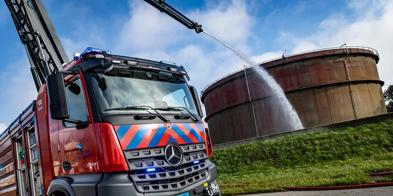 Feuerwehrfahrzeug von Mercedes-Benz mit ausfahrbarem Leiter, das Wasser auf einen großen, zylindrischen Tank spritzt. Hintergründe zeigen einen klaren Himmel und Grasflächen.
