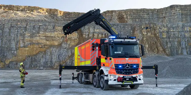 Ein Feuerwehrfahrzeug von Mercedes-Benz mit Kranaufsatz hebt eine Containerbox in einem Steinbruch. Ein Feuerwehrmann steht in sicherem Abstand und beobachtet die Aktion.