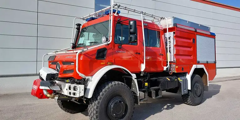 Ein rotes Feuerwehrfahrzeug von Mercedes-Benz mit einem robusten Design und einer Lkw-Kabine steht vor einer modernen Wand. Das Fahrzeug ist für den Einsatz im Gelände ausgelegt.