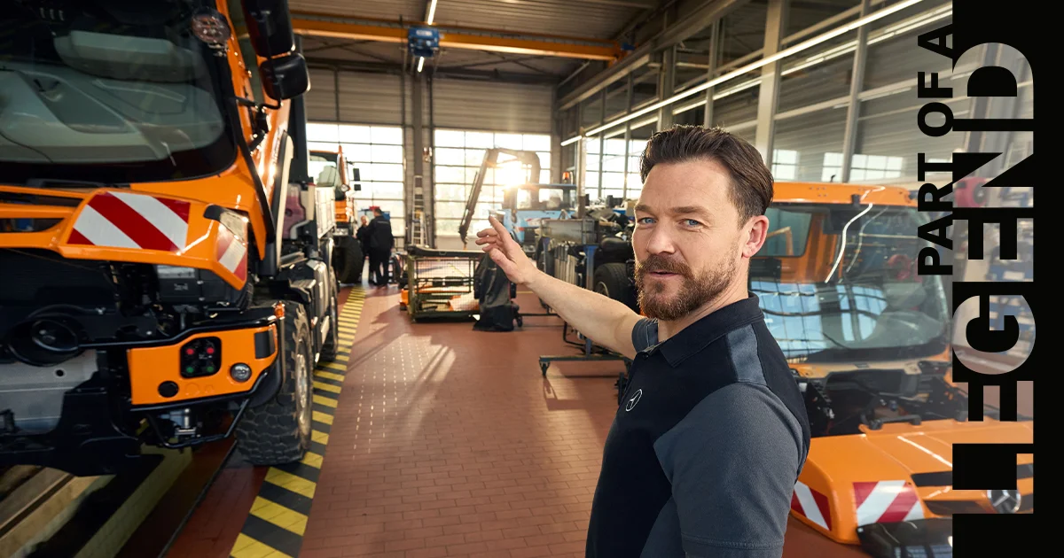 A smiling technician wearing a safety jacket is posing in an auto repair shop, with a mechanic working in the background. Text overlay reads "PART OF A LEGEND.