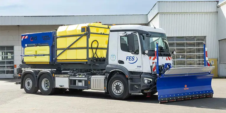 Ein Mercedes-Benz Lkw mit einem blauen Schneepflug und einem gelben Tanksystem steht auf einem Firmengelände, bereit für den Einsatz im Winterdienst.