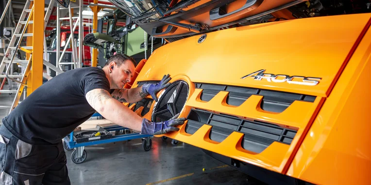 Un operaio sta installando il logo di Mercedes-Benz su un camion Arcos di colore arancione, mentre lavora nella fase di assemblaggio in un'officina.