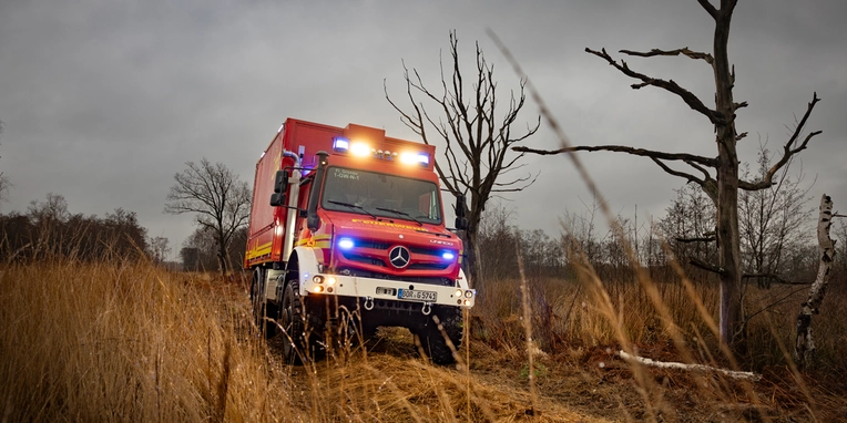 Ein Mercedes-Benz Feuerwehrfahrzeug mit Blaulicht fährt durch hohes, trockenes Gras in einer ländlichen Umgebung, umgeben von kahlen Bäumen und bewölktem Himmel.