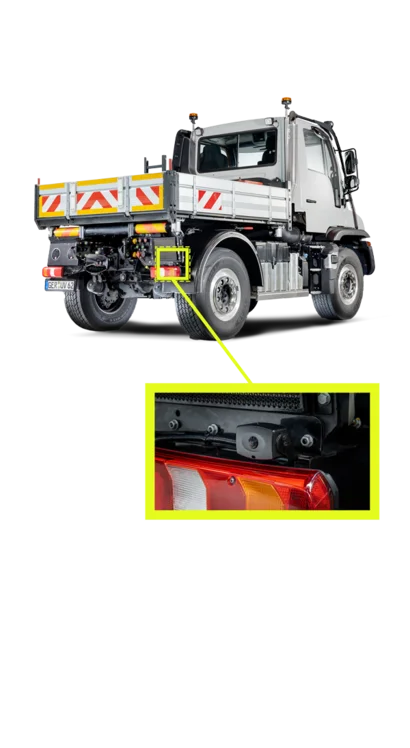 Ein Unimog von Mercedes-Benz, der von hinten zu sehen ist. Die Rückansicht zeigt die Ladefläche mit Warnmarkierungen und einem Detailbereich, der ein Rücklicht und die Befestigung zeigt.