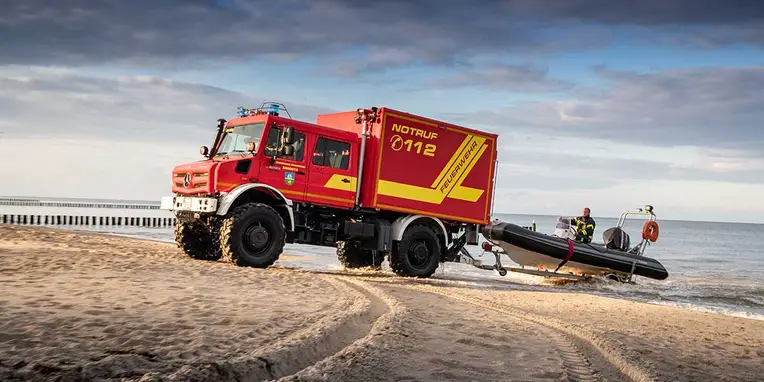 Ein rotes Feuerwehrfahrzeug von Mercedes-Benz fährt am Strand und zieht ein Schlauchboot ins Wasser, während zwei Retter sich auf dem Boot vorbereiten.