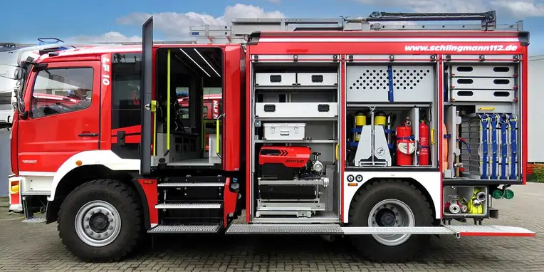 Rotes Feuerwehrfahrzeug mit aufklappbaren Seitenabteilen, ausgestattet mit Schläuchen, Feuerlöschern und Werkzeugen. Sichtbar sind verschiedene Einsatzgeräte für den Brandschutz und die Rettung.