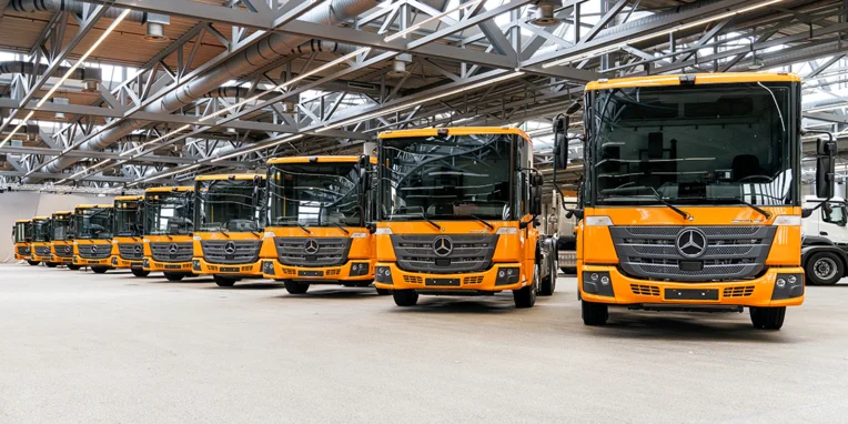 Eine Reihe orangefarbener Mercedes-Benz Lkw steht in einer modernen Lagerhalle aufgereiht, bereit für den Einsatz.