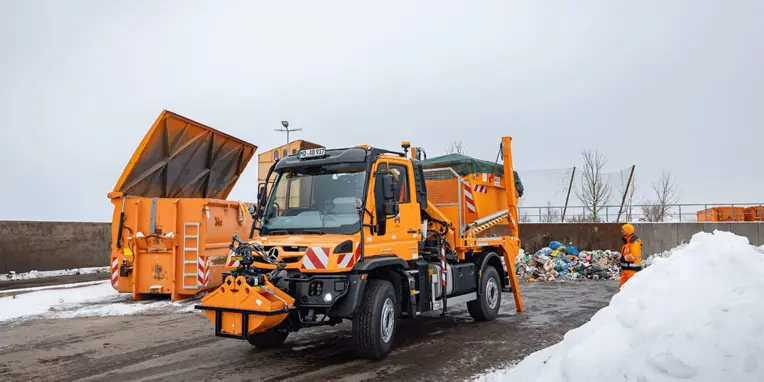 Ein orangefarbener Mercedes-Benz Müllwagen entleert einen Abfallcontainer an einer Recyclingstelle, während ein Mitarbeiter mit leuchtender Sicherheitskleidung den Müllsortierbereich überwacht.