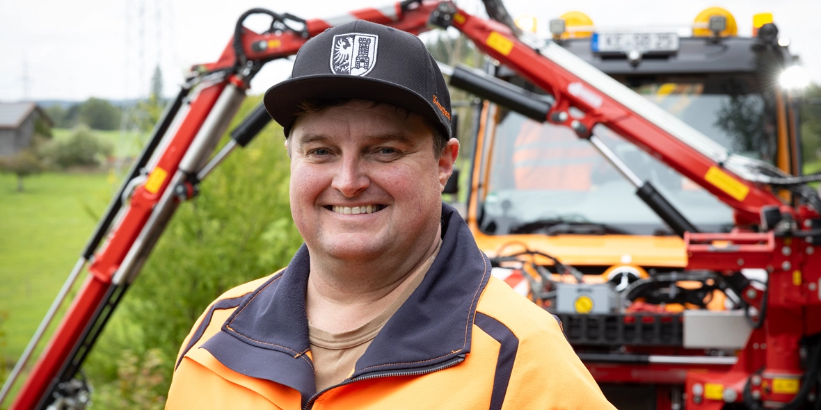 Ein lächelnder Mann in orangefarbener Arbeitskleidung steht vor einem roten Arbeitsfahrzeug mit Auslegern. Das Bild vermittelt eine positive Arbeitsatmosphäre im Freien.