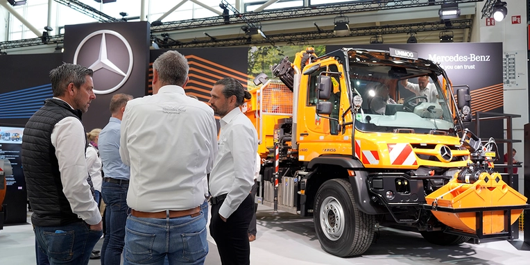 Fachbesucher stehen an einem Messestand von Mercedes-Benz und betrachten einen gelben Nutzfahrzeug. Gespräche über Fahrzeugmerkmale und Einsatzmöglichkeiten finden statt.