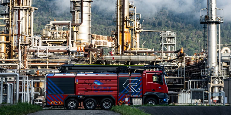 Ein roter Mercedes-Benz-Lkw steht vor einer Raffinerie mit großen, metallischen Anlagen und Rohrleitungen. Die Szene ist klar und zeigt industrielle Aktivität in einer industriellen Umgebung.