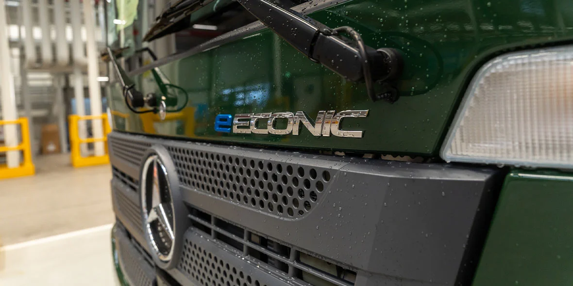 Nahaufnahme des vorderen Bereichs eines grünen Mercedes E-Conic Fahrzeugs. Das Logo und der Schriftzug "eCONIC" sind deutlich sichtbar. Die Oberfläche ist leicht nass, was auf eine kürzliche Reinigung oder Regen hindeutet. Im Hintergrund sind unscharfe industrielle Elemente zu erkennen.