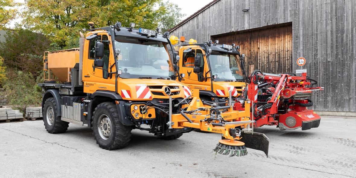Zwei gelbe Mercedes-Benz Nutzfahrzeuge stehen in einer Auffahrt, eines mit einer Kehrmaschine vorne. Im Hintergrund ist ein Holzgebäude sichtbar.