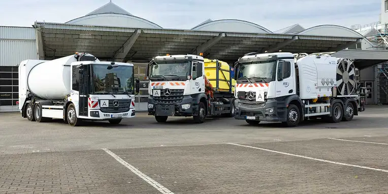 Drei Mercedes-Benz Nutzfahrzeuge stehen auf einem Parkplatz. Das erste Fahrzeug ist ein Zementmischer, gefolgt von einem Müllfahrzeug und einem großen Lkw mit einem Kesselaufbau.