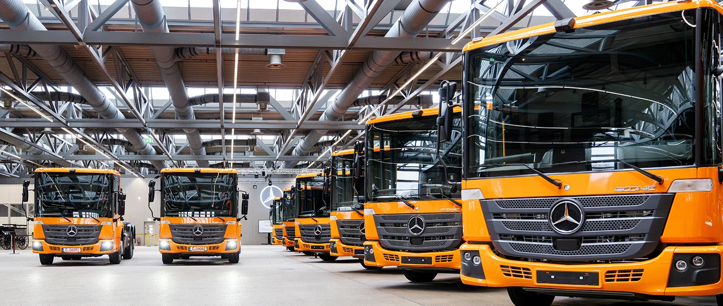 Sechs orangefarbene Mercedes-Benz-Busse stehen in einer modernen Halle, gut ausgeleuchtet und ordentlich aufgereiht, bereit für den Einsatz oder die Inspektion.