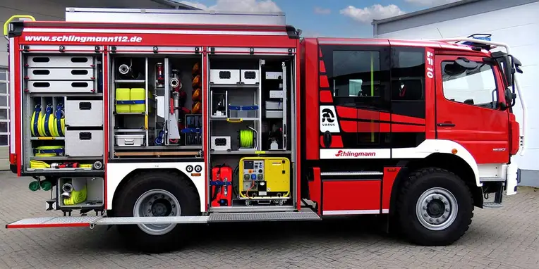 Ein rotes Feuerwehrfahrzeug mit offenen Seitenablagen, die verschiedene Feuerwehrgeräte und -ausrüstungen wie Schläuche, Kästen und Werkzeuge präsentieren.