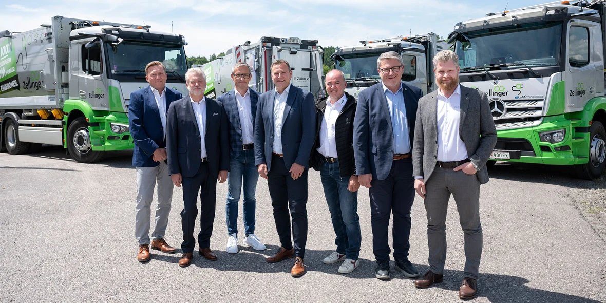 Grupo de siete hombres posando frente a camiones eActros de Mercedes-Benz, diseñados para la recolección de residuos, con un fondo despejado. Todos llevan trajes formales y algunos muestran sonrisas, simbolizando la colaboración en el sector de la logística sostenible.