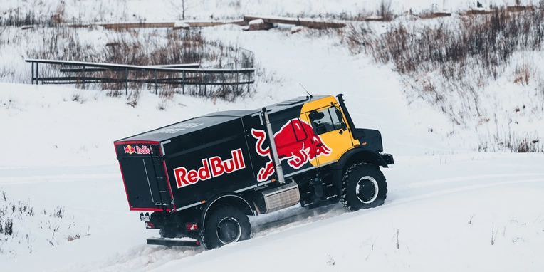 Ein offroad-tauglicher Lkw mit Red Bull-Design fährt über verschneite Hügel. Der Lkw ist robust und geeignet für winterliche Bedingungen.