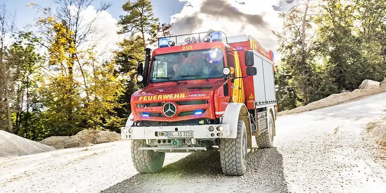 Ein rotes Feuerwehrfahrzeug von Mercedes-Benz fährt auf einer unbefestigten Straße, umgeben von Bäumen und natürlicher Landschaft. Blaulichter sind aktiviert.
