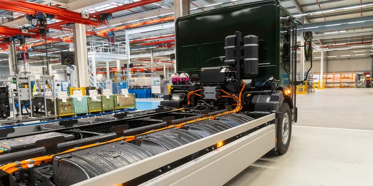 Ein Lkw mit sichtbar ausgelegter Technik steht in einer modernen Produktionshalle. Die Rückansicht zeigt verschiedene Komponenten, darunter Tankstellen und Kabel, auf einer offenen Fahrgestellbauweise. Umgebende Produktionsanlagen sind angedeutet, jedoch nicht im Fokus.