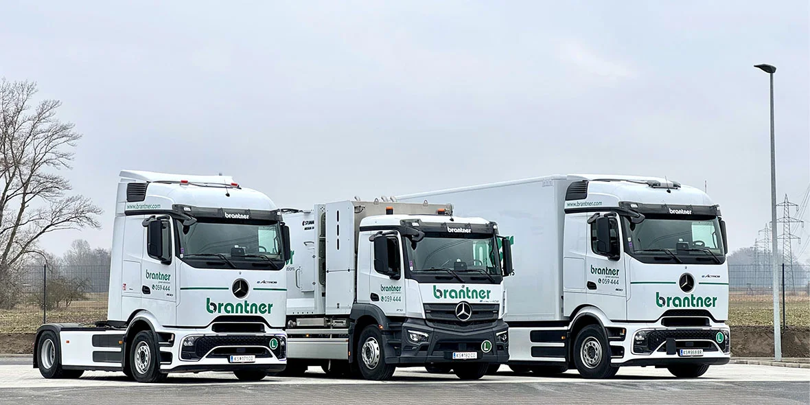 Drei Mercedes-Benz Lkw stehen nebeneinander auf einem Parkplatz. Alle Fahrzeuge sind in Weiß mit grünen Schriftzügen der Firma Brantner lackiert.