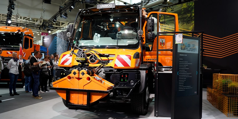 Ein orangefarbener Mercedes-Benz-Lkw mit Schneeschild wird auf einer Messe präsentiert. Menschen stehen im Hintergrund und betrachten das Fahrzeug.