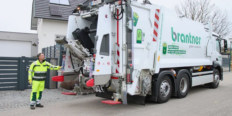 Ein Müllwagen vor einem Wohnhaus, mit einem Mitarbeiter in reflektierender Kleidung, der den Ladevorgang für Mülltonnen überwacht. Auf der Seite des Wagens steht das Logo von branther.