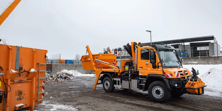 Ein orangefarbener Mercedes-Benz Unimog steht auf einem schneebedeckten Gelände neben einem Abfallcontainer. Im Hintergrund sind Müll und Recyclingmaterialien sichtbar.